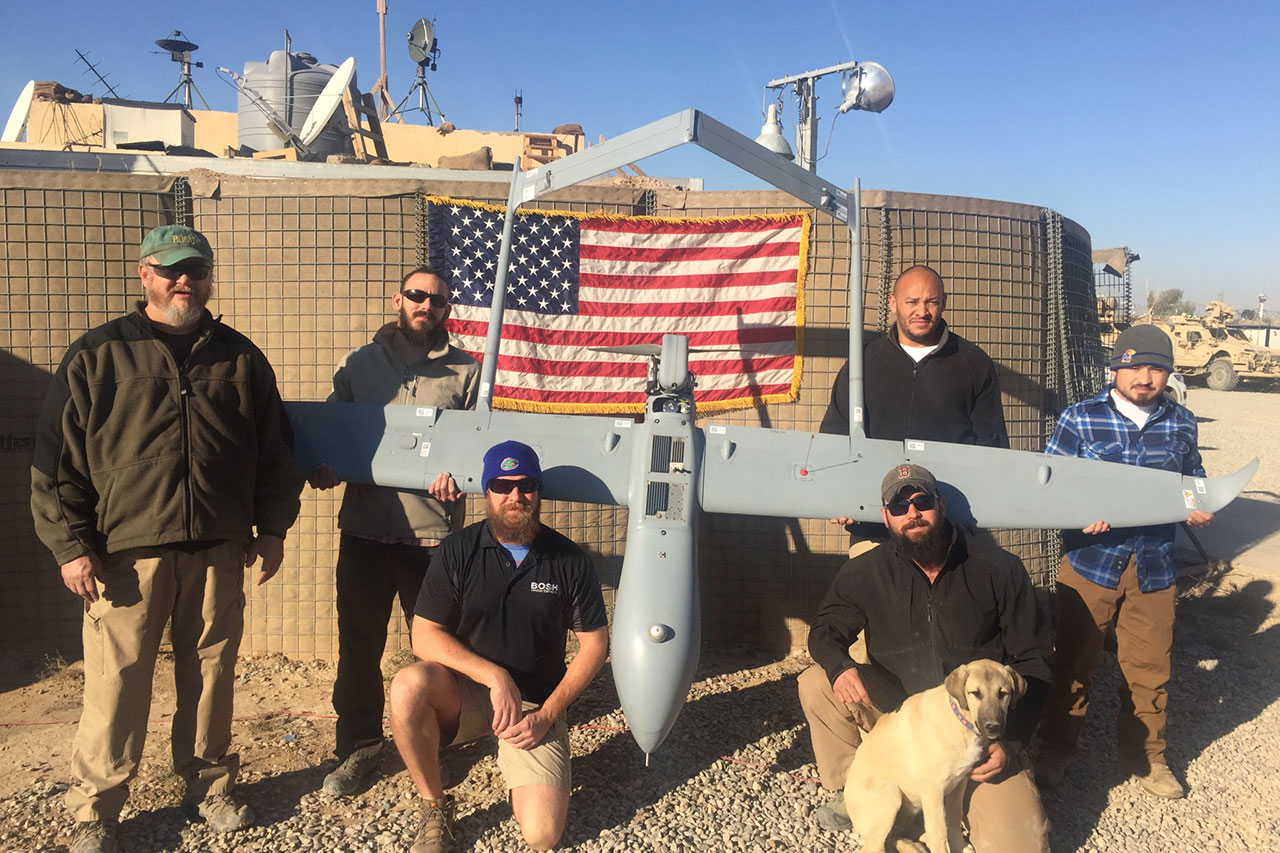 Six men posing around a drone. The drone is being held up in front of an American Flag