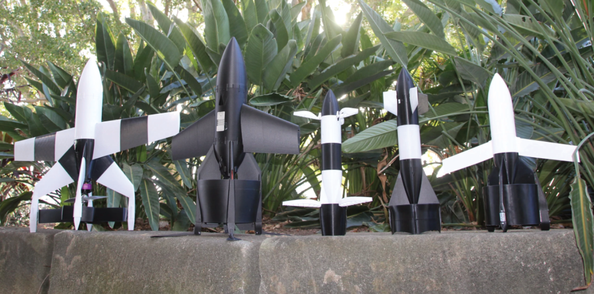 Five handmade black-and-white paper rocket models lined up on a concrete wall with green tropical plants in the background.
