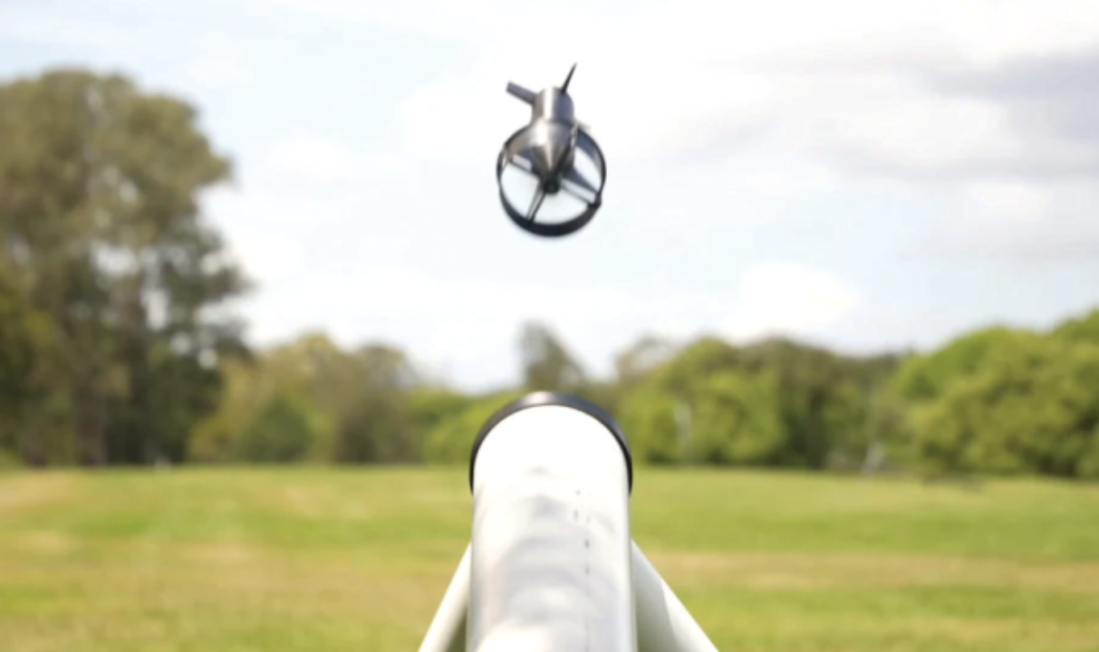 Small metallic drone arcing upward from a white tube-style launcher in a grassy field.