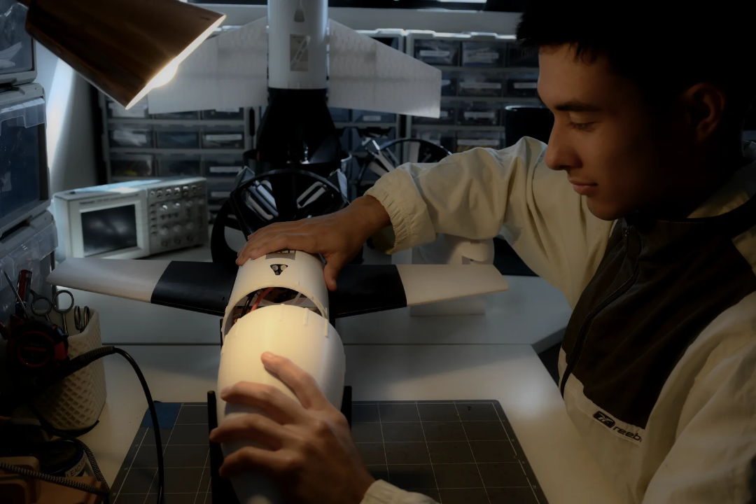 Person in a lab coat works on a model airplane fuselage at a cluttered workstation with tools nearby.